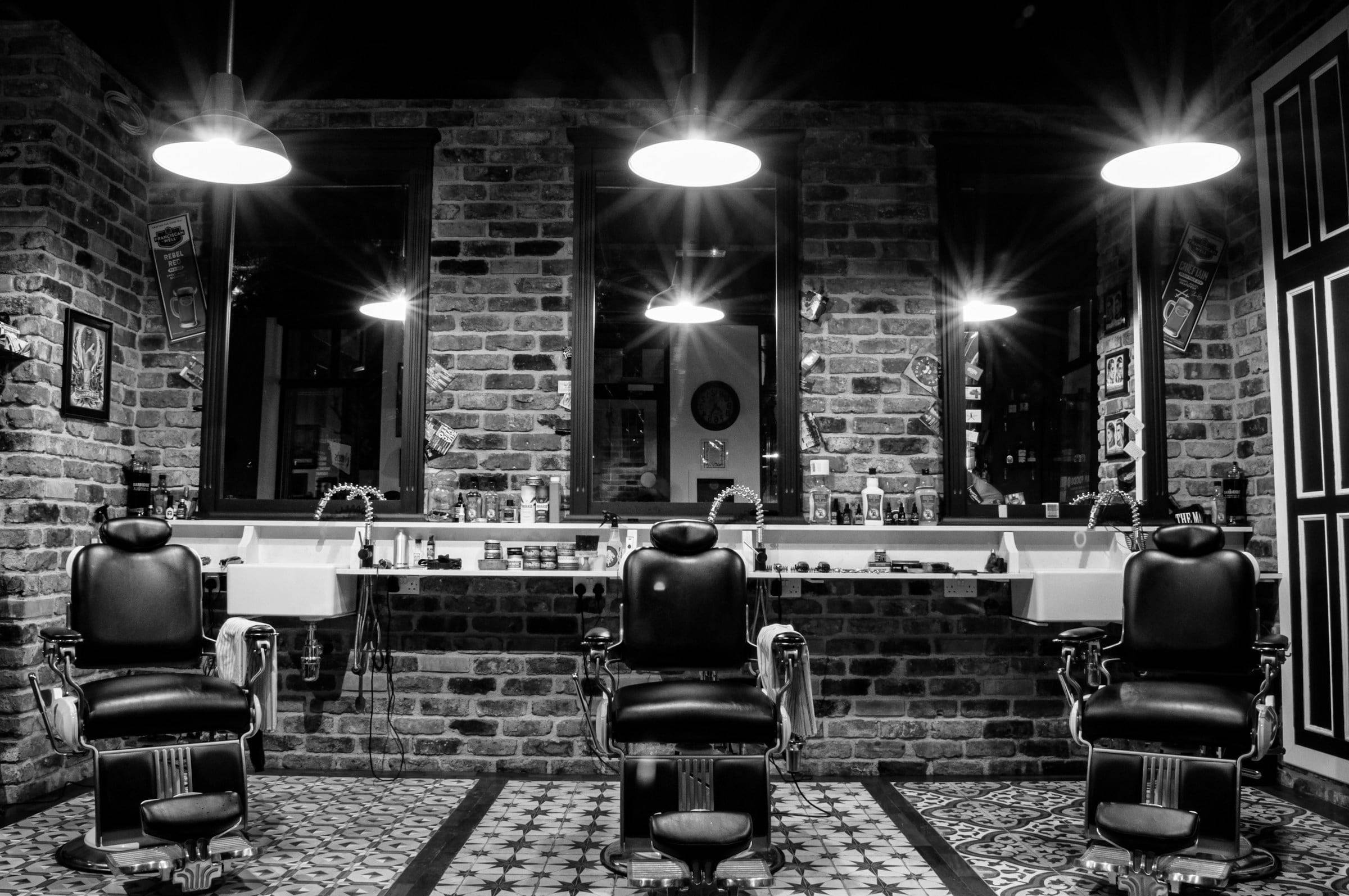 Dunkler Salon mit Backsteinwand, drei Barberstühlen und Pendelleuchten